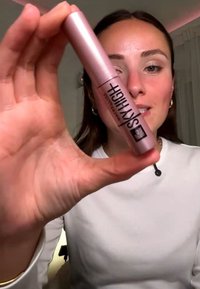 Vrouw houdt een roze Maybelline Sky High-mascarabuizen dicht bij de camera, draagt een wit topje en gouden oorringen.