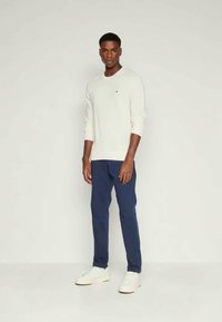 Helles Cremeshirt mit langen Ärmeln, dunkelblaue Hose und weiße Turnschuhe. Das Shirt hat ein kleines Logo auf der Brust.