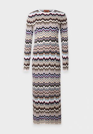 Un mannequin est debout devant un fond uni clair, portant une robe longue aux manches longues avec des rayures en zigzag multicolores et des sandales noires.
