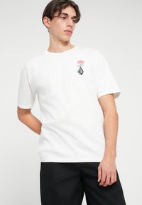 T-shirt en coton blanc avec un graphique de rose et une forme géométrique noire sur la poitrine. Manches courtes et col rond.