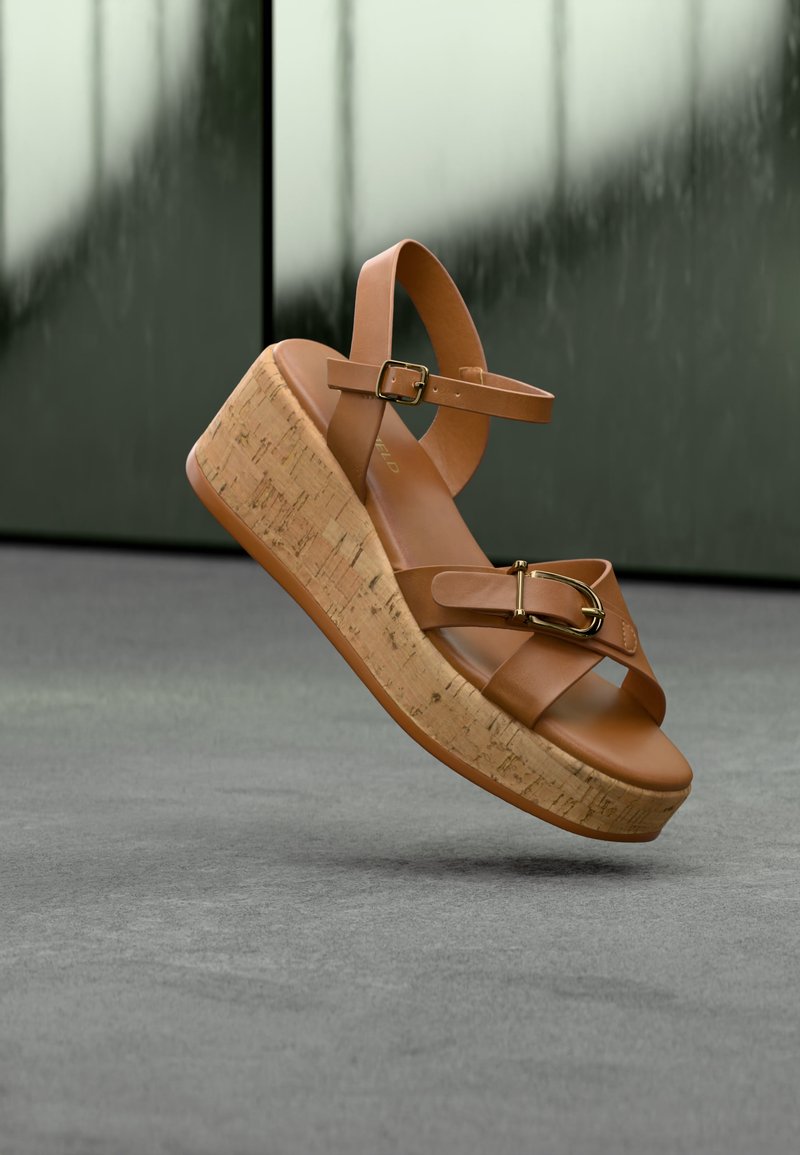 Καφέ δερμάτινο πλατφόρμα σάνδαλο με φελλό πάτο, λουρί στον αστράγαλο με αγκράφα και χρυσή λεπτομέρεια αγκράφας στα μπροστινά λουριά, εμφανίζεται σε θολό φόντο.