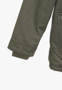 Rękaw oliwkowej kurtki z prążkowanym ściągaczem i widocznymi detalami przeszycia w pobliżu kieszeni i dołu.