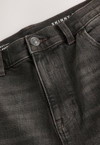 Jean en denim noir avec fermeture par bouton, passants de ceinture et étiquette « SKINNY » visible à l'intérieur de la ceinture.