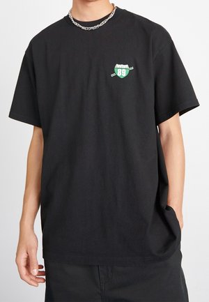 Mężczyzna noszący czarny, oversize t-shirt z zielono-białym emblematem "WIP 89" na piersi i srebrnym łańcuszkiem, ręka w kieszeni.