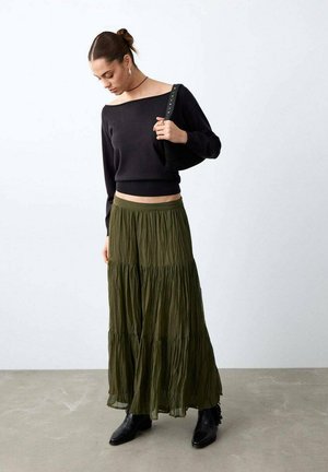 Woman wearing off-shoulder black sweater, olive green tiered maxi skirt, black boots, and carrying a black studded shoulder bag, looking down.