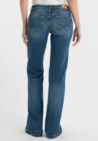 Jean bootcut en denim bleu moyen, avec une taille haute, deux poches arrière ornées d'un motif de couture distinctif et une texture légèrement usée.