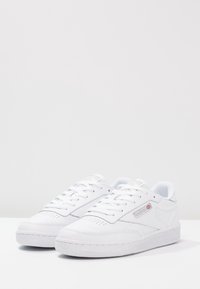 Chaussures de sport blanches en cuir lisse, dotées d'un embout perforé, de lacets plats et d'une semelle en caoutchouc texturée. Accentuées par un marquage discret.