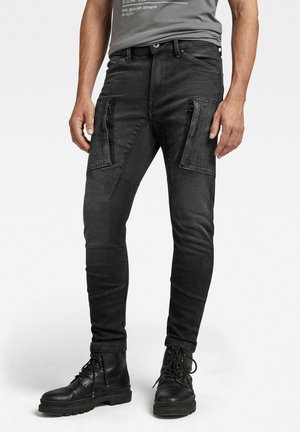 Mężczyzna w czarnych, dopasowanych spodniach cargo z kieszeniami na udach zapinanymi na zamek oraz czarnych sznurowanych butach, stoi z rękami swobodnie ułożonymi po bokach.