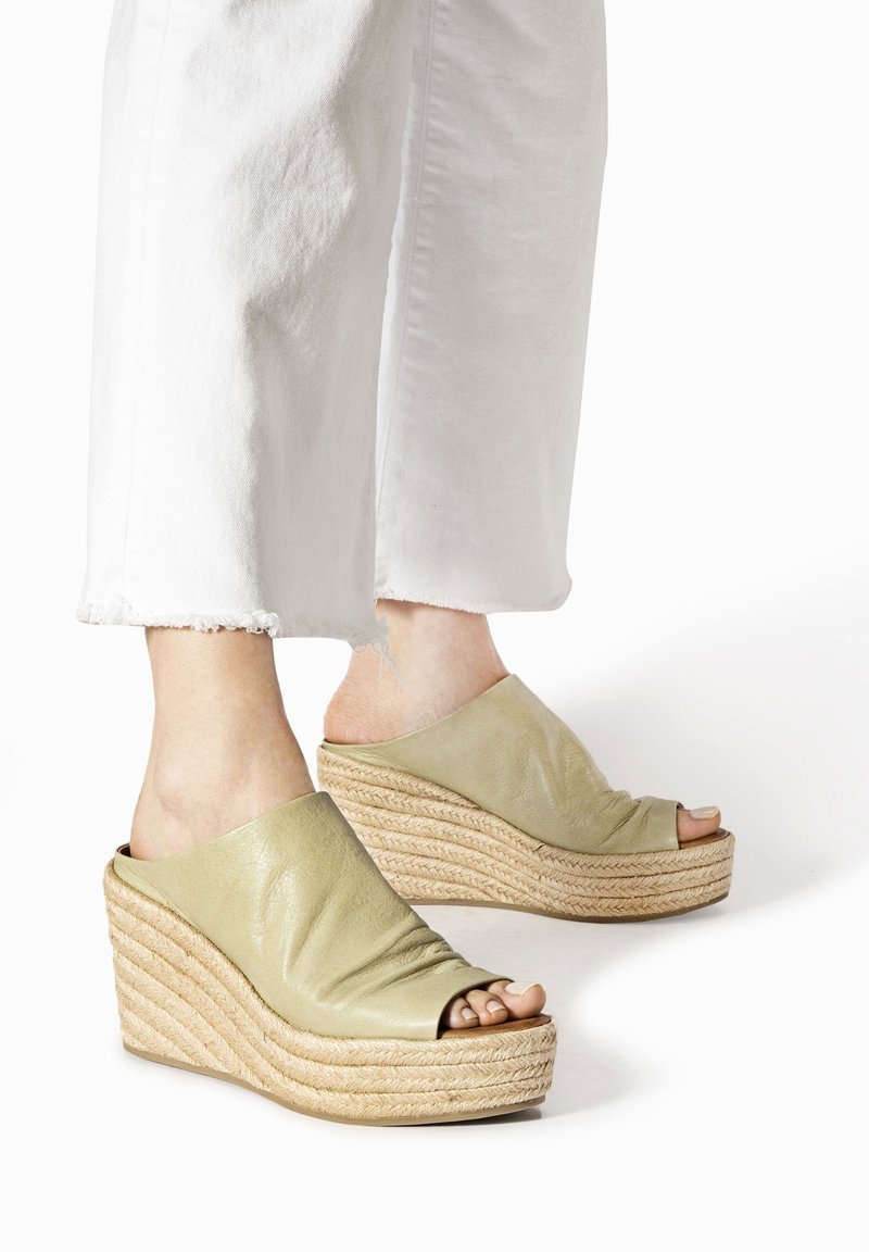 Pieds portant des sandales compensées en cuir vert clair à bout ouvert, associées à un pantalon blanc court à ourlet effiloché, sur fond blanc.