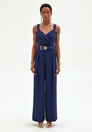 Donna in piedi che indossa una tuta senza maniche blu navy con cintura, pantaloni ampi, orecchini dorati e sandali rigati con punta aperta.