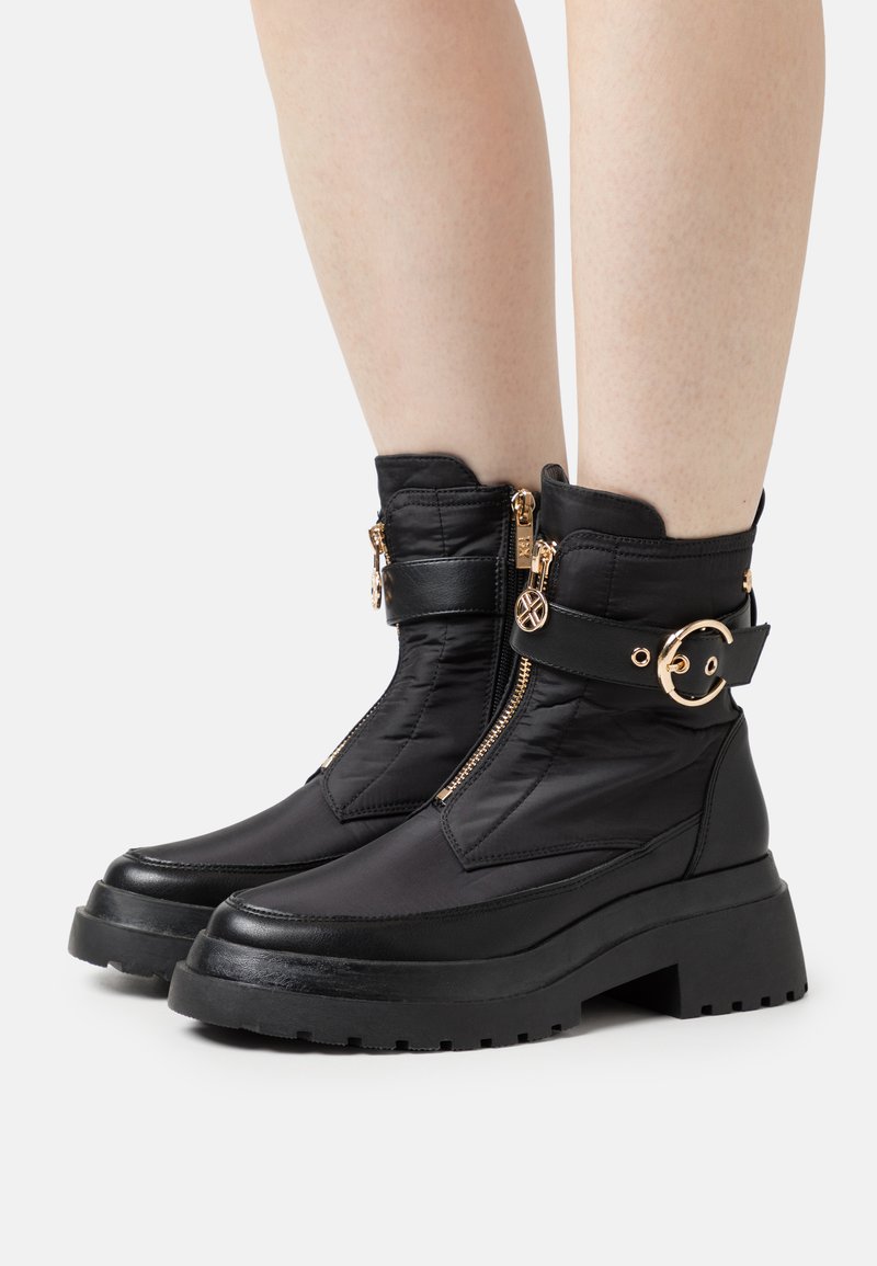 Botas de tobillo negras con material sintético texturizado, suela gruesa, detalle del zipper dorado y una correa decorativa con herraje circular.