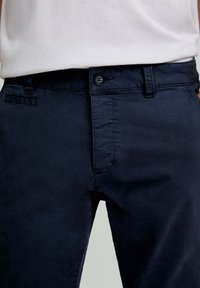 Marineblauwe katoenen chino's met een knoopsluiting, voorzakken en een gladde textuur. De tailleband heeft riemlussen voor een verstelbare pasvorm.