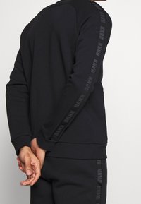 Persoon in zwart sweatshirt en broek met herhaalde "KNVB" logo strepen op de mouwen en zijkanten, handen samengevoegd achter de rug.