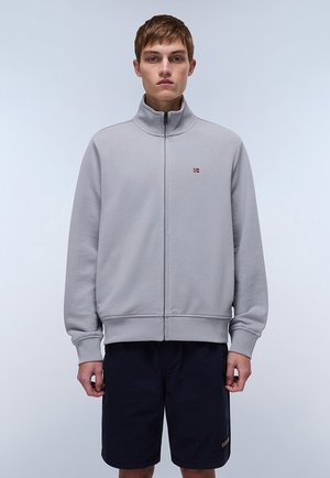 Siva zip-up jopica z visokim ovratnikom in rebrastimi manšetami, nosena čez temne kratke hlače. Ima majhno logo na levem prsnem predelu.
