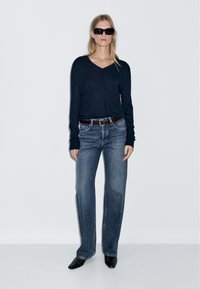 Navy long-sleeve V-neck top, blue loose-fit jeans, brown belt, black heeled shoes, and oversized black sunglasses. Smooth textures and casual fit.