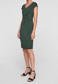 Grön knälång klänning med V-ringning, korta ärmar och en vriden frontdesign. Bärs med svarta sandaler med hög klack och remmar. Slät textur.
