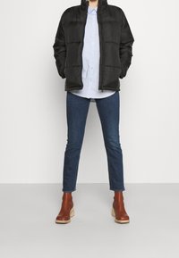 Persona vestida con una chaqueta acolchada negra, una camisa de botones azul claro, jeans azul oscuro y botas de tobillo con tacón marrones, de pie sobre un fondo sencillo.