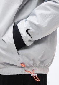 Λευκό μανίκι μπουφάν Nike με μαύρο μανσέτι, το χέρι του φορέα στην τσέπη, και πορτοκαλί κορδόνι με πλαστικό κούμπωμα στο τελείωμα.