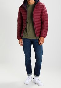 Bordeaux puffer jakke med en marineblå hætte, grøn mønster T-shirt, blå jeans og hvide sneakers med sorte accenter.