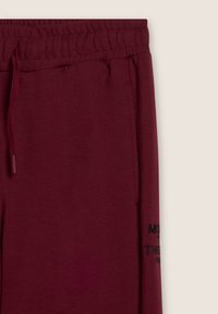 Pantaloni della tuta bordeaux con cintura elastica e cordoncino. Include tasche laterali e testo ricamato in nero sulla gamba sinistra. Textura morbida.
