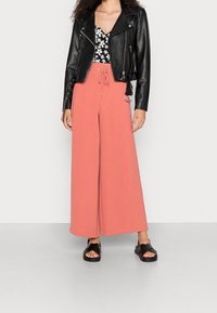 Svart läderbikerjacka över en svart och vit blommig topp, kombinerad med vida korallbyxor och svarta sandaler. Släta texturer och avslappnad passform.