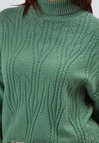 Grüner Strickpullover mit hohem Rippkragen, der ein strukturiertes Muster aus Zopfmuster und Rauten aufweist. Weicher Stoff, lockere Passform.