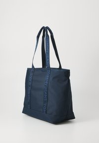 Geantă de umăr bleumarin dintr-un material durabil, cu două mânere lungi ce poartă marca "TOMMY JEANS", formă rectangulară și textură fină.