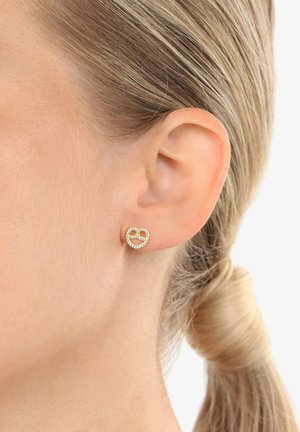 Nahaufnahme eines Frauenohrs, das einen kleinen, goldenen Ohrstecker in Form von Brezeln mit eingelegten klaren Steinen trägt, während das blonde Haar zurückgebunden ist.