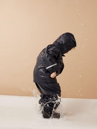 Zwarte waterdichte jas en broek met elastische manchetten, capuchon en reflecterende accenten. Kind spettert in het water op een lichte achtergrond.