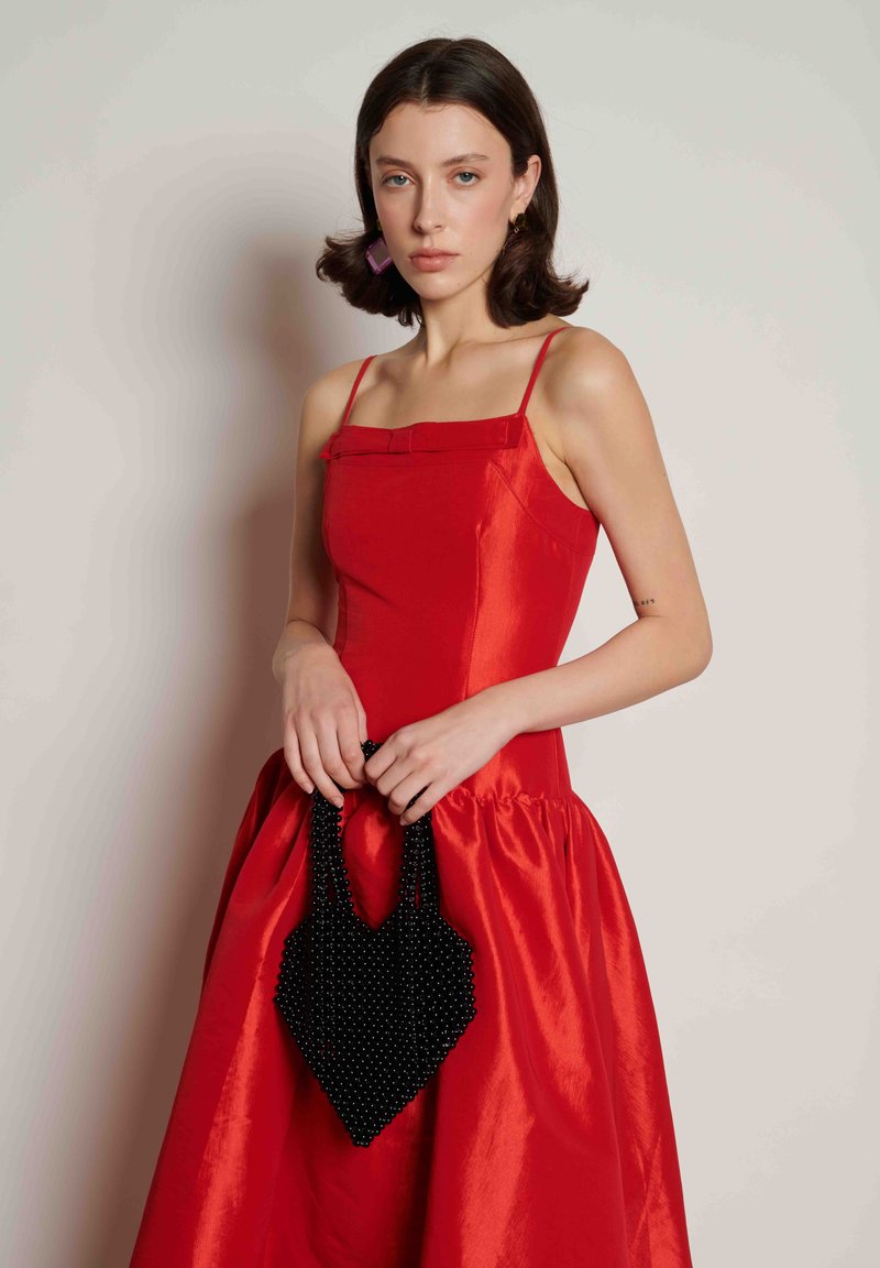 Vestido de cetim vermelho com corpete estruturado, detalhe de laço e saia rodada, combinado com uma mala preta em forma de coração decorada com contas. Textura suave, cor viva.