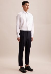 Camicia bianca a maniche lunghe con bottoni e tasca, abbinata a pantaloni scuri su misura e scarpe oxford nere. Design semplice e moderno.
