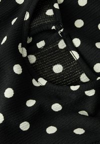 Zwarte ribstof met grote witte stippen en een close-up van een ronde halslijn met een zwarte knop op een gevouwen kledingstuk.