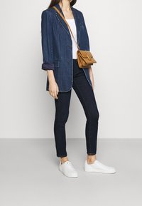 Blazer de mezclilla oscura con mangas enrolladas, camiseta blanca sin mangas, jeans skinny oscuros y zapatillas blancas. Bolso cruzado de color marrón claro con detalle de solapa.