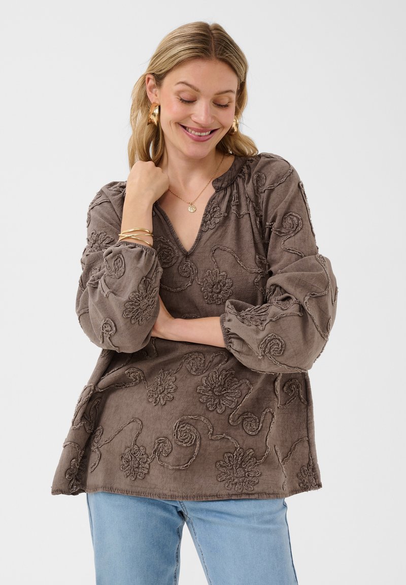 Femme aux cheveux clairs portant un chemisier marron à motifs floraux texturés à manches longues et un jean bleu clair, souriante avec une main près du cou.