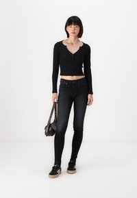 Černý dlouhý rukáv zkrácený top s detaily na knoflíky, doplněný o tmavé skinny džíny. Modelka nese malou hnědou tašku a má na sobě černé tenisky.
