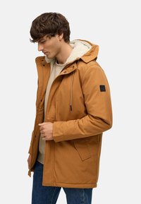 Brūna bieza jaka ar kapuci, ar šerpa oderi, priekšējo rāvējslēdzēju, spiedpogām un melnu logo marķējumu uz kreisā piedurknes.