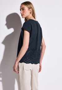 Top blu navy con maniche corte, caratterizzato da un orlo a scalloped e da intricati motivi ritagliati nella parte inferiore. Materiale liscio con una vestibilità comoda.