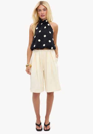 Mujer rubia con top negro sin mangas tipo halter con lunares blancos, pantalones cortos anchos color crema hasta la rodilla, sandalias negras y una pulsera dorada.