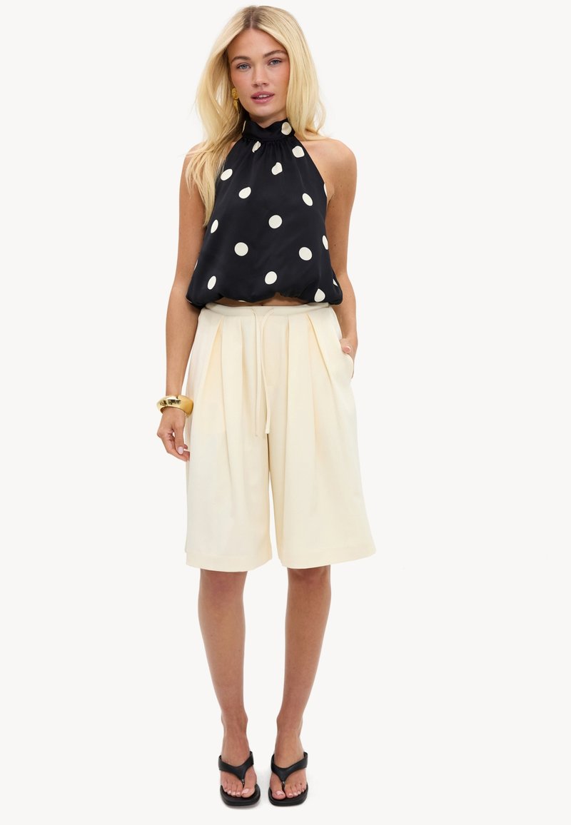 Femme blonde portant un haut sans manches noir à pois blancs, un short large crème longueur genoux, des sandales noires et un bracelet en or.