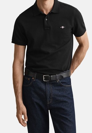 Mand iført en tætsiddende sort polo-shirt med lille logo på brystet, mørkeblå jeans og et sort bælte med en sølv spænde.