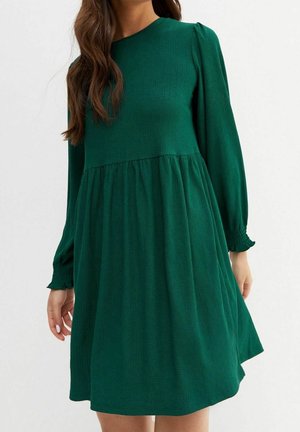 Kvinde iført en langærmet, mørkegrøn, tekstureret knælængde kjole med indsnævret talje og rynkede ærmekanter.