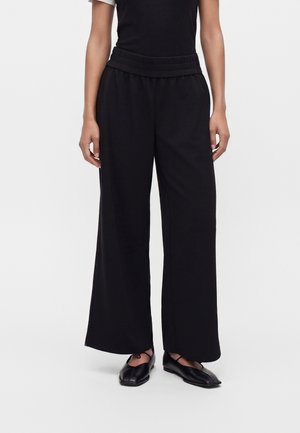 Person wearing black wide-leg trousers with a high elastic waistband and black flat shoes with crisscross straps, standing against a plain background.