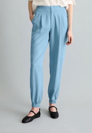 Vrouw draagt lichtblauwe elastische broek met omgeslagen pijpen, gecombineerd met zwarte Mary Jane schoenen en een wit ingestopt overhemd.