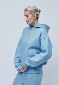 Hanorac albastru deschis, cu o croială lejeră, glugă mare și manșete riboudate. Prezintă o broderie simplă a logo-ului pe piept. Fabrică moale și textură netedă.