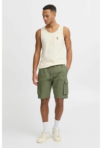 Top bej fără mâneci cu un buzunar, pantaloni scurți cargo verzi cu buzunare laterale, șosete albe și adidași crem. Ținută casual cu croială lejeră.