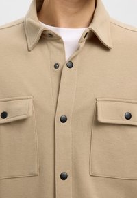 Veste beige à boutons pression noirs avec deux poches avant à rabat portée sur une chemise blanche.