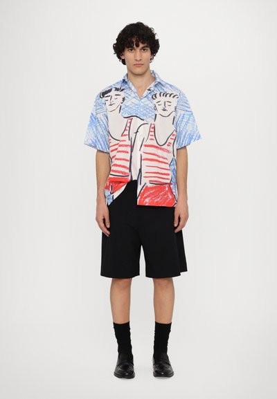 Camisa de manga corta con estampado gráfico azul y rojo de figuras, combinada con pantalones cortos negros hasta la rodilla y zapatos y calcetines negros.