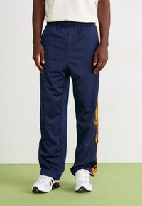 Person med marineblå Adidas træningsbukser med orange striber og hvide sneakers, der står på et lysegrønt gulv foran en ensfarvet væg.