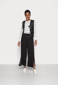 Gilet en cuir noir sur une chemise à rayures blanches et noires à manches longues, associé à un pantalon noir ample avec des fentes sur les côtés et des baskets blanches.