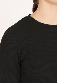 Schwarzes, geripptes Langarmoberteil mit rundem Ausschnitt. Der Stoff hat eine weiche, strukturierte Optik und schmiegt sich eng an den Körper, ohne sichtbare Nähte.